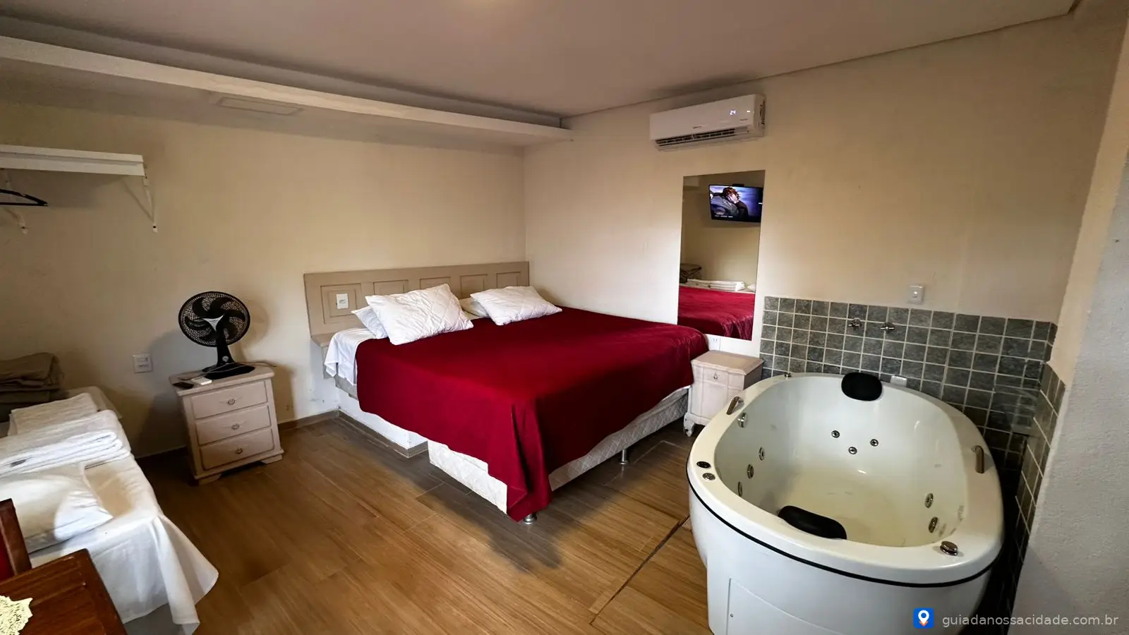 Quarto com cama de casal com colcha vermelha, banheira de hidromassagem e ar-condicionado na parede.